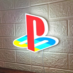 Puede incluir: Una luz de logotipo de PlayStation blanca, azul, amarilla y roja. La luz tiene la forma del logotipo de PlayStation y est&aacute; iluminada con luz blanca.