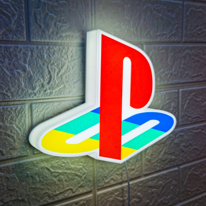 Puede incluir: Una luz de logotipo de PlayStation blanca, roja, amarilla, azul y verde. El logotipo est&aacute; iluminado y est&aacute; montado en una pared.