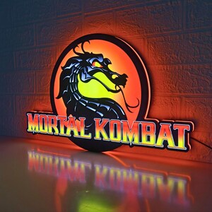 K&ouml;nnte beinhalten: Beleuchtetes Mortal Kombat-Schild mit einer schwarzen Drachensilhouette auf einem kreisf&ouml;rmigen orange-gelben Hintergrund. Die Worte "MORTAL KOMBAT" sind in einer fetten, leuchtenden Schrift dargestellt. Das Schild ist an einer Backsteinmauer befestigt.