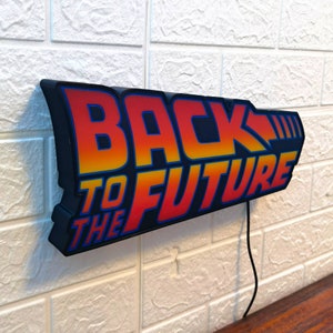 Op de afbeelding: Een zwart "Back to the Future" bord met de tekst in een oranje en gele gradi&euml;nt. Het bord is bevestigd aan een witte bakstenen muur en heeft een zwart netsnoer.