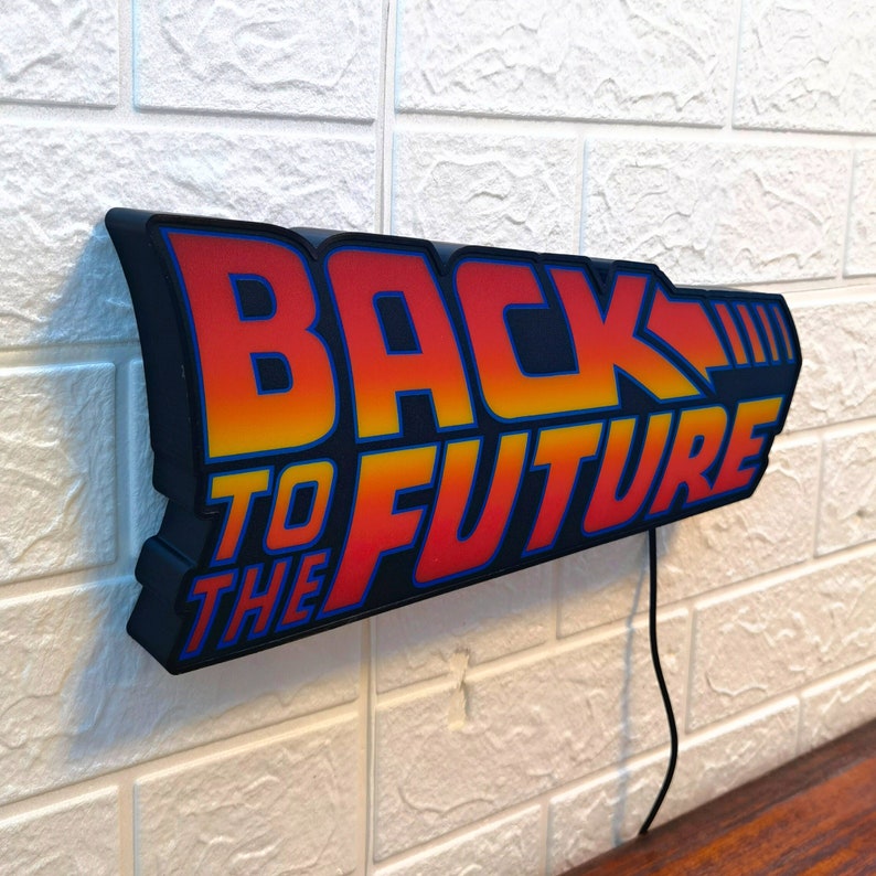 Op de afbeelding: Een zwart "Back to the Future" bord met de tekst in een oranje en gele gradi&euml;nt. Het bord is bevestigd aan een witte bakstenen muur en heeft een zwart netsnoer.