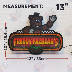 May include: A Freddy Fazbear's Pizza Place sign featuring a brown bear character waving. The sign measures 13 inches wide by 10 inches tall. The text reads "FREDDY FAZBEAR'S Pizza PLACE" in yellow and green lettering against a dark background.