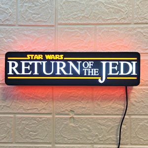 Puede incluir: Un letrero rectangular negro con las palabras "STAR WARS RETURN OF THE JEDI" en letras blancas con un contorno amarillo. El letrero está iluminado con luces rojas y blancas.