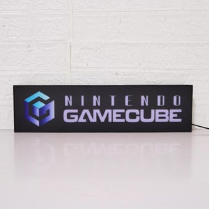 May include: A black rectangular sign with the Nintendo GameCube logo in the upper left corner and the text "NINTENDO GAMECUBE" in white letters.