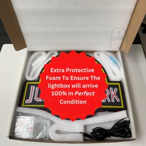 May include: A rectangular Jurassic Park lightbox packaged inside a cardboard box with white protective foam inserts. The lightbox features black, red, and yellow colors with white lettering. A black power cord and small hardware bag are included.