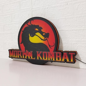 K&ouml;nnte beinhalten: Beleuchtetes Mortal Kombat-Schild mit dem Logo des Spiels: eine schwarze Drachensilhouette vor rotem und gelbem Hintergrund, darunter die Worte "MORTAL KOMBAT". Das Schild ist an einer wei&szlig;en Backsteinmauer befestigt.