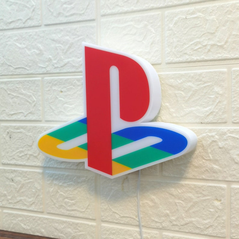 Puede incluir: Una luz de pared con el logotipo de PlayStation en blanco, rojo, azul, verde y amarillo. El logotipo es una letra P estilizada con una l&iacute;nea curva que se extiende desde la parte inferior.