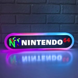 Puede incluir: Un letrero rectangular iluminado con el logotipo de Nintendo 64. El letrero tiene un fondo negro con las palabras "NINTENDO 64" en blanco y rojo. El logotipo presenta una "N" colorida y un diseño 3D. El letrero está rodeado por un degradado de rosa, azul y morado.