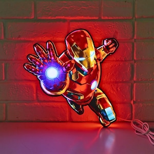 May include: A red and gold Iron Man LED light in a dynamic pose, with a glowing blue energy blast emanating from his hand. The superhero figure is mounted on a brick wall, with a white cord and plug visible.