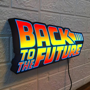Op de afbeelding: Een verlicht bord met de tekst "BACK TO THE FUTURE" in een gestileerd lettertype. Het bord heeft een zwarte rand met een verloop van rood, oranje en geel, omlijnd in blauw. Het is bevestigd aan een witte bakstenen muur.