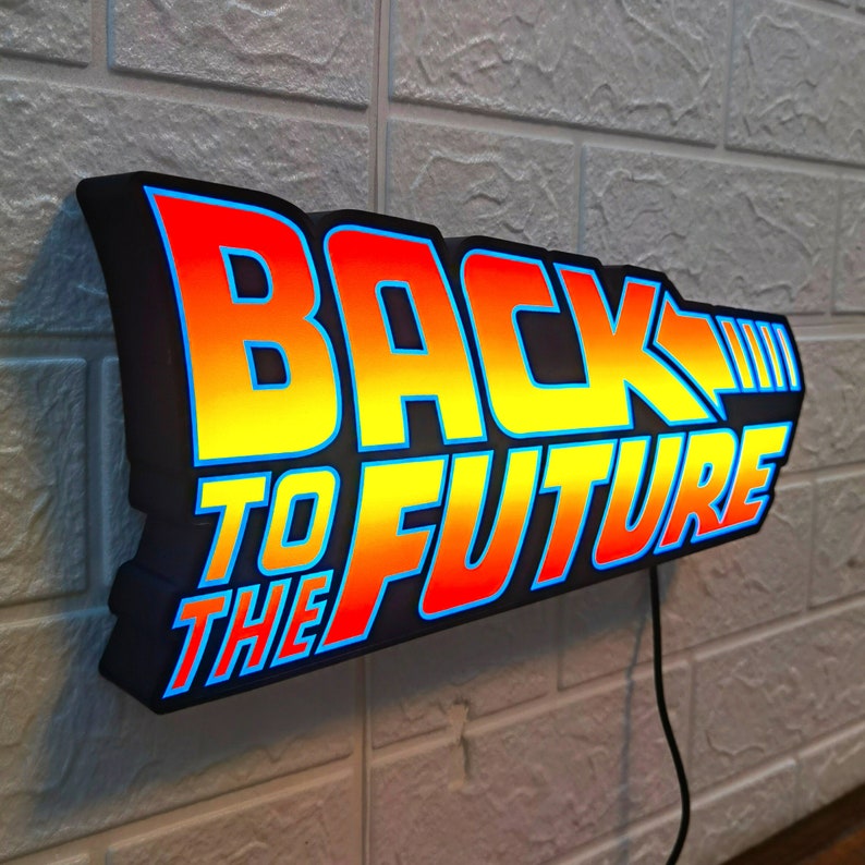 Op de afbeelding: Een verlicht bord met de tekst "BACK TO THE FUTURE" in een gestileerd lettertype. Het bord heeft een zwarte rand met een verloop van rood, oranje en geel, omlijnd in blauw. Het is bevestigd aan een witte bakstenen muur.
