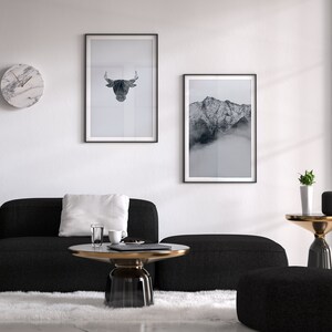 May include: A black sectional sofa with a white pillow, a round glass coffee table with a gold rim, a black and gold side table with a potted plant, and two framed black and white prints on a white wall.