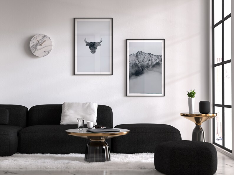 May include: A black sectional sofa with a white pillow, a round glass coffee table with a gold rim, a black and gold side table with a potted plant, and two framed black and white prints on a white wall.