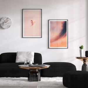 May include: A living room interior with a black sectional sofa, a white rug, a round glass coffee table, a black and gold side table, a potted plant, and two framed prints on the wall. One print features a bird in flight against a pink background, and the other print features a sunset sky.