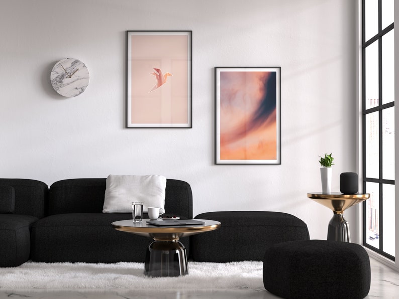 May include: A living room interior with a black sectional sofa, a white rug, a round glass coffee table, a black and gold side table, a potted plant, and two framed prints on the wall. One print features a bird in flight against a pink background, and the other print features a sunset sky.