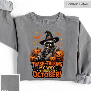 May include: A grey crewneck sweatshirt featuring a graphic of a raccoon wearing a witch hat and sunglasses, holding a cup. The raccoon is surrounded by three jack-o'-lanterns, bats, and stars. The text reads, "TRASH IS MY SPIRIT ANIMAL" and "TRASH-TALKING MY WAY THROUGH OCTOBER!"