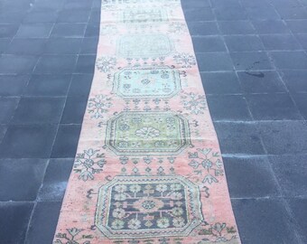 Puede incluir: Una alfombra de pasillo larga y estrecha con una base de color rosa coral desvanecido. La alfombra muestra un patrón repetitivo de cuatro medallones octogonales en tonos verde salvia, azul verdoso y carbón, con motivos florales y geométricos.