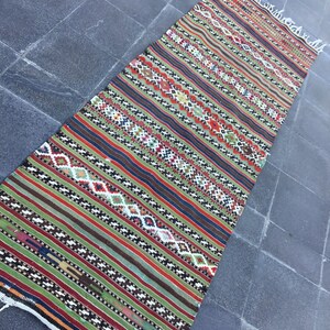 May include: A long, rectangular rug with a striped pattern in shades of green, red, brown, and blue. The rug features geometric diamond shapes and fringe on the ends. This handmade rug is suitable for a hallway or entryway.