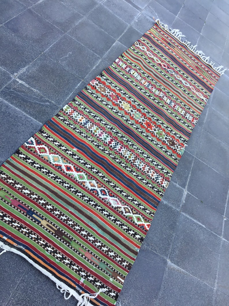 May include: A long, rectangular rug with a striped pattern in shades of green, red, brown, and blue. The rug features geometric diamond shapes and fringe on the ends. This handmade rug is suitable for a hallway or entryway.