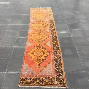 Pode incluir: Um tapete longo e estreito com um design persa tradicional. O tapete é principalmente laranja e amarelo com detalhes em preto e marrom. O tapete tem franjas em ambas as extremidades.