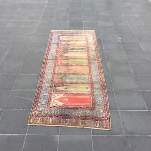 May include: Rectangular, vintage rug with a detailed border and central design. The rug features a red, green, and beige color palette with intricate patterns and text. The rug is placed on a gray tiled surface.