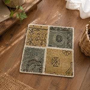 May include: A square patchwork rug with a cream-colored border. The rug is divided into four sections, each with a different pattern in shades of olive green, gray, and black. The rug is placed on a wooden floor.