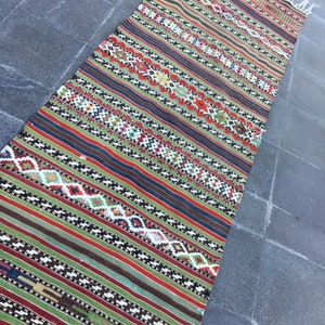 May include: A rectangular, hand-woven rug with a vibrant, striped design. The rug features a mix of colors including green, red, blue, and brown, with intricate geometric patterns. The rug has a fringed edge and is likely made of natural fibers.
