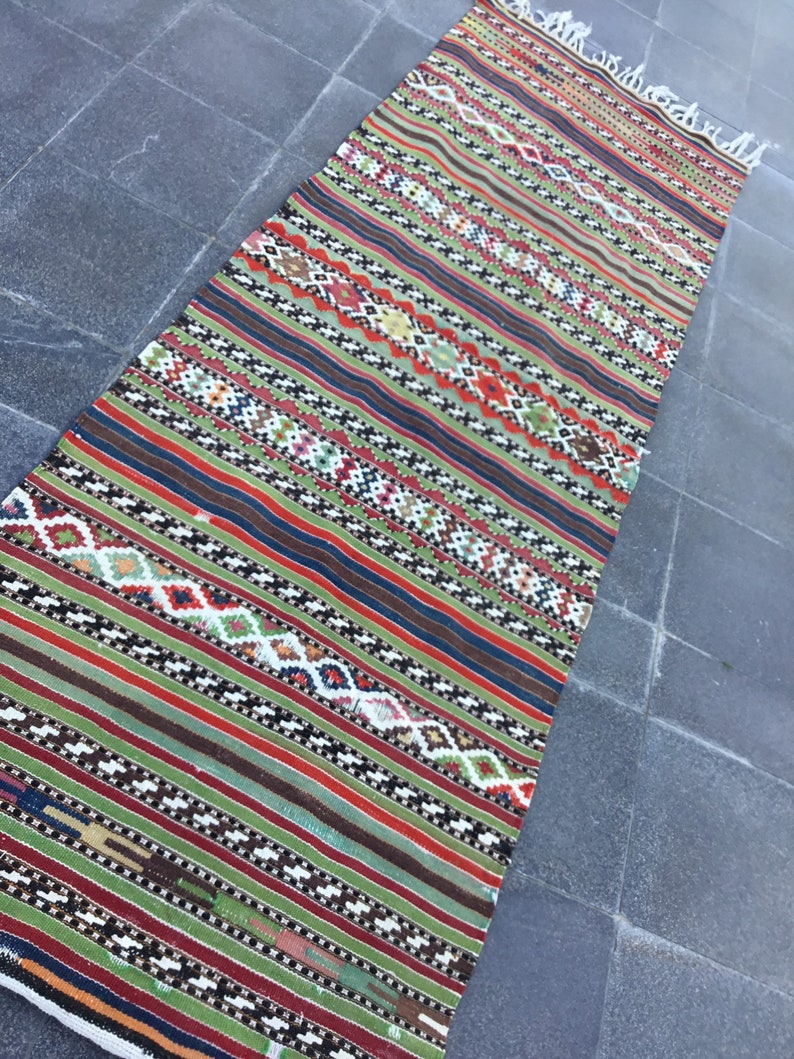 May include: A rectangular, hand-woven rug with a vibrant, striped design. The rug features a mix of colors including green, red, blue, and brown, with intricate geometric patterns. The rug has a fringed edge and is likely made of natural fibers.