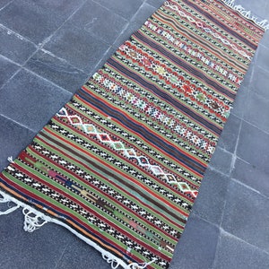 May include: A rectangular, handwoven rug with a colorful, striped pattern. The rug features a mix of red, green, brown, and blue stripes, with diamond and geometric designs. The edges have white fringe.