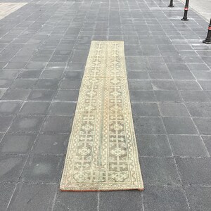 以下が含まれることがあります： ベージュの背景に、茶色、緑、白の幾何学模様が施された長方形の敷物。敷物は灰色のタイル張りの床の上に置かれている。