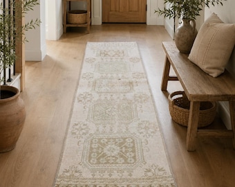 May include: A rectangular, cream-colored runner rug with a traditional, geometric pattern in shades of green and tan. The rug has a distressed look and a fringed edge, suitable for a hallway or entryway.