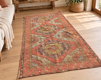 Puede incluir: Una alfombra rectangular con un patrón geométrico repetitivo en tonos naranja terracota, verde salvia y morado oscuro. La alfombra presenta una fila central de medallones en forma de diamante con motivos florales y un borde.