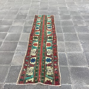 以下が含まれることがあります： 中央に赤いストライプ、緑とクリーム色のセクションを配した長方形の模様入りラグ。赤、青、緑で繰り返される花柄と幾何学模様が特徴です。ラグは灰色のタイルの上に置かれています。