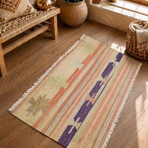 May include: A rectangular area rug with a geometric pattern in shades of beige, orange, purple, and green. The rug has a striped design with a fringed edge, suitable for a living room or bedroom.