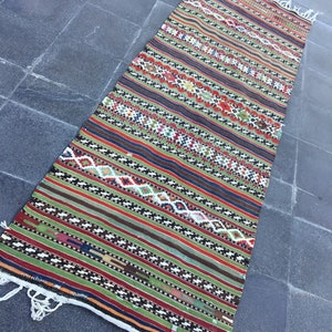 May include: A long, rectangular rug with a vibrant, striped design. The rug features a mix of colors including red, green, brown, and white, with geometric patterns. The edges have white fringe. This rug is suitable for a hallway or entryway.