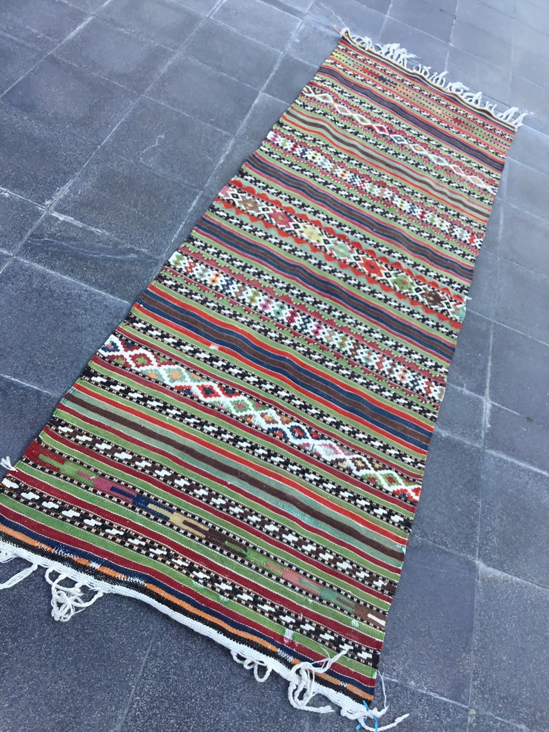 May include: A long, rectangular rug with a vibrant, striped design. The rug features a mix of colors including red, green, brown, and white, with geometric patterns. The edges have white fringe. This rug is suitable for a hallway or entryway.