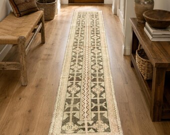 May include: A long, narrow runner rug featuring a repeating geometric pattern in dark olive green and cream. The rug has a central vertical zigzag border and is finished with short, vibrant red fringe along the narrow ends of the rectangular textile.