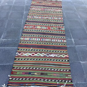 May include: A long, rectangular rug with a vibrant, striped pattern. The rug features alternating bands of color, including red, green, and blue, with geometric designs. White fringe accents the ends. This rug is suitable for a hallway or entryway.