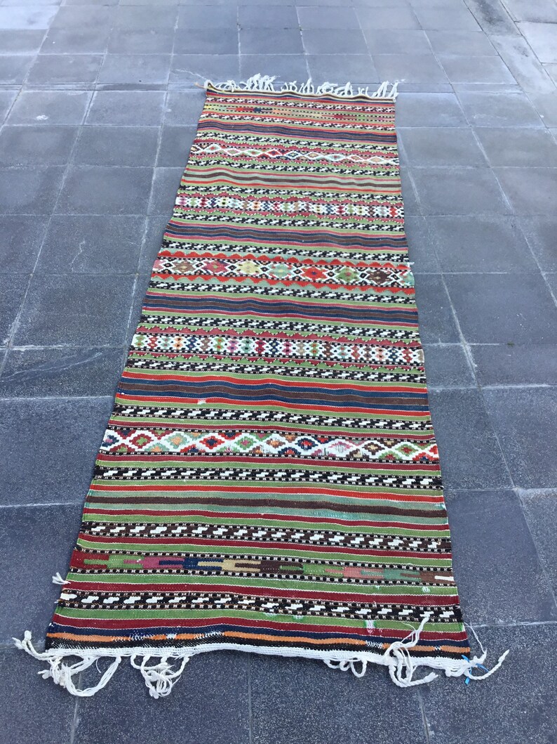 May include: A long, rectangular rug with a vibrant, striped pattern. The rug features alternating bands of color, including red, green, and blue, with geometric designs. White fringe accents the ends. This rug is suitable for a hallway or entryway.