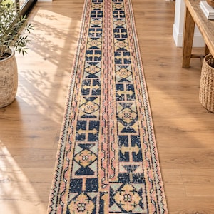 May include: A long, narrow runner rug featuring a repeating geometric pattern in navy blue, soft pink, cream, and muted yellow. The design consists of rectangular panels with intricate borders and central floral-inspired motifs throughout.