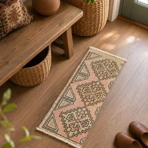 Könnte beinhalten: Ein rechteckiger Teppich mit rosa Hintergrund und geometrischen Mustern in Beige, Grün und Schwarz. Der Teppich liegt auf einem Holzboden, in der Nähe einer Bank und eines Weidenkorbs. Braune Lederslipper sind ebenfalls zu sehen.