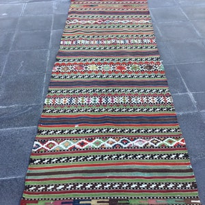 May include: A long, rectangular rug with a striped pattern in various colors, including red, green, and blue. The rug has a woven texture and a fringe on both ends. The design features geometric shapes and horizontal bands.