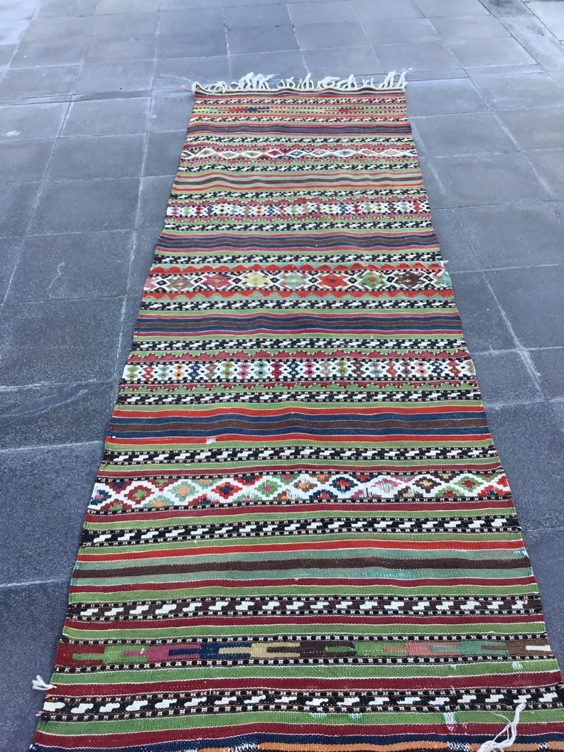 May include: A long, rectangular rug with a striped pattern in various colors, including red, green, and blue. The rug has a woven texture and a fringe on both ends. The design features geometric shapes and horizontal bands.