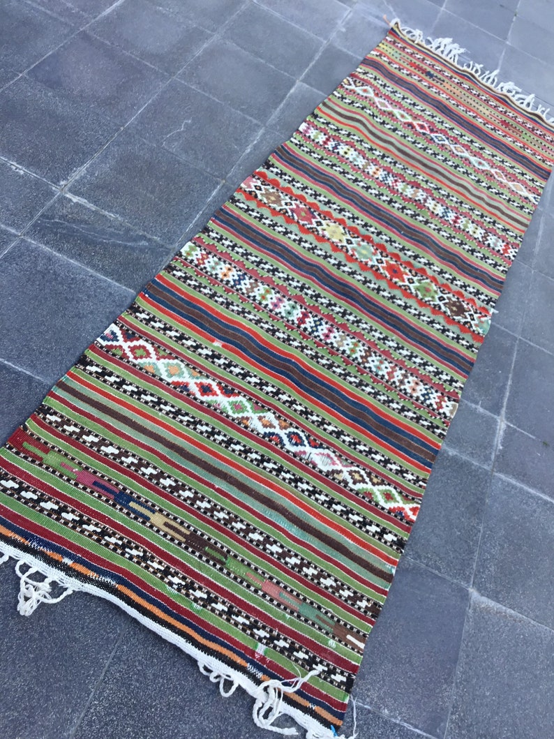 May include: A long, rectangular rug with a vibrant, geometric pattern. The rug features alternating stripes of green, red, brown, and blue, with intricate diamond and zigzag designs. White fringe edges the rug's ends, adding a textural contrast.