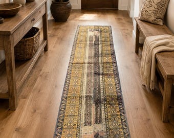 May include: A long, rectangular runner rug featuring a complex geometric pattern in shades of mustard yellow, charcoal gray, and faded pink. The rug has a dark border with repeating motifs and a central panel with intricate, symmetrical designs.