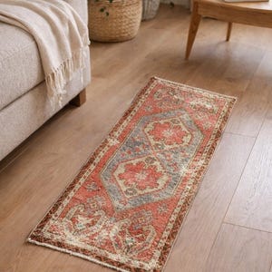 Puede incluir: Una alfombra rectangular de estilo vintage con un aspecto desgastado. La alfombra presenta un diseño central en tonos rojos, azules y beige, con un borde marrón oscuro. La alfombra está colocada sobre un suelo de madera.