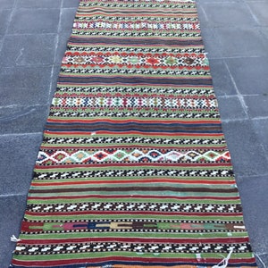 May include: A long, rectangular rug with a vibrant, striped design. The rug features a mix of colors, including red, green, blue, and white, with geometric patterns and fringe on the ends. The rug is likely made of woven fabric.