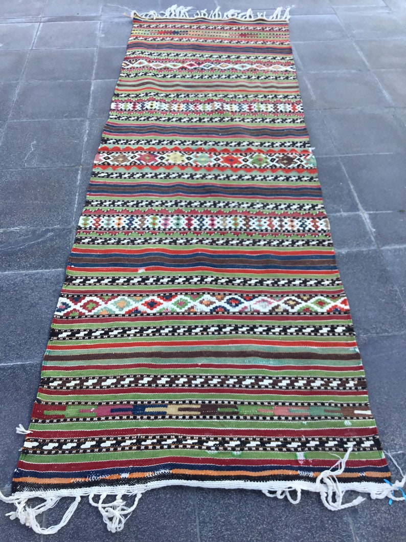 May include: A long, rectangular rug with a vibrant, striped design. The rug features a mix of colors, including red, green, blue, and white, with geometric patterns and fringe on the ends. The rug is likely made of woven fabric.
