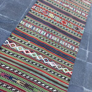 May include: A colorful, rectangular rug with a striped pattern. The rug features alternating bands of green, red, brown, and blue, with intricate geometric designs in white, black, and other colors. The rug is made of woven fabric.