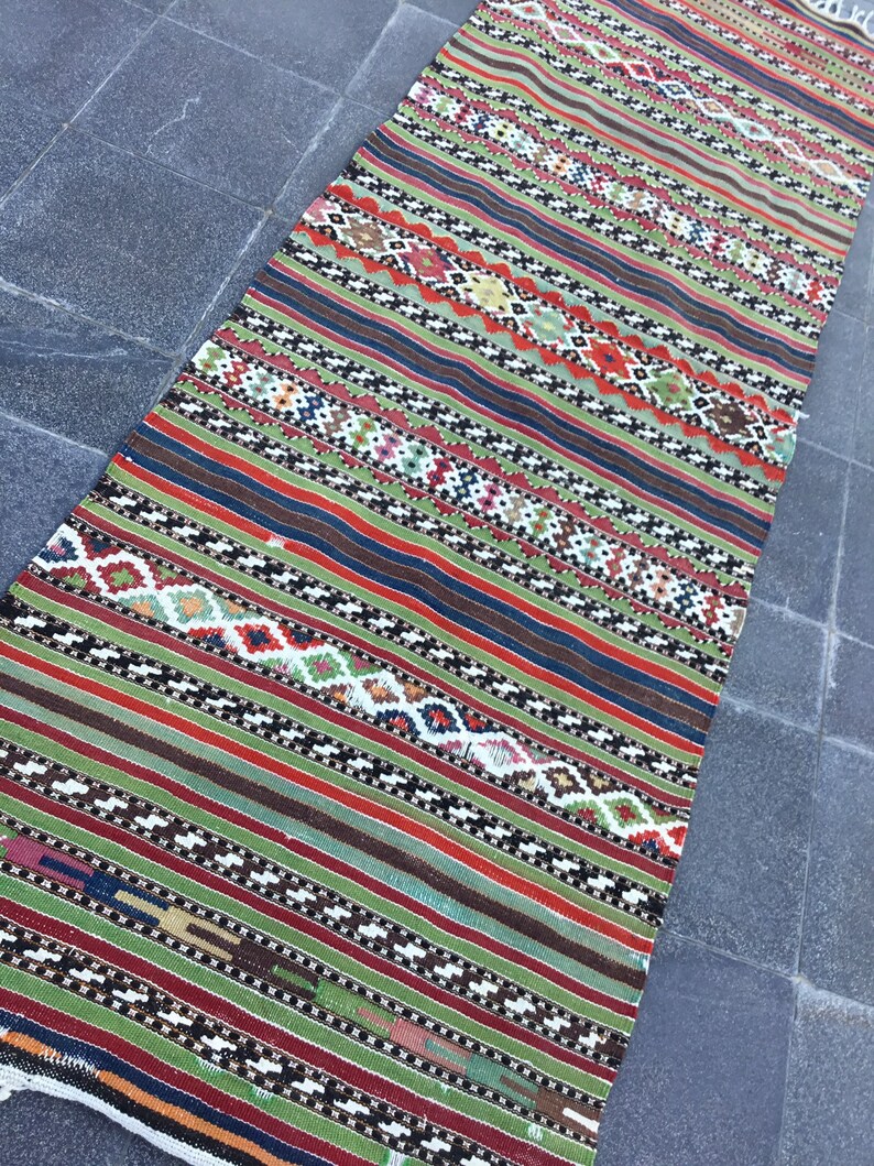 May include: A colorful, rectangular rug with a striped pattern. The rug features alternating bands of green, red, brown, and blue, with intricate geometric designs in white, black, and other colors. The rug is made of woven fabric.